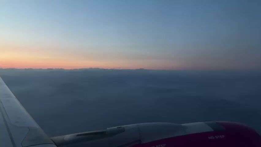 The video shows a view from an airplane window during dawn or dusk. The sky is a gradient of soft blues and pinks near the horizon. Below the sky, a layer of dark clouds or mist obscures the landscape, which appears to be mountainous. The wing of the airplane, with its white leading edge and pink accent, is visible in the foreground, along with part of an engine nacelle. There are some markings on the engine nacelle, including the words "NO STEP". The camera is stationary.