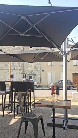 The video shows an outdoor seating area with several tables and chairs. Large black umbrellas are providing shade. Two wine glasses sit on a small table. In the background, there is a street with a car and buildings. The scene appears to be a cafe or restaurant patio.