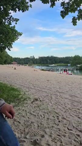 The video shows a sandy beach next to a body of water. Several people are scattered along the beach and in the water. Some are sitting on the sand, while others appear to be swimming or wading. There are trees and foliage surrounding the beach and water. The sky is partly cloudy with a blue hue. In the distance, there are some structures, possibly piers or docks, extending into the water, and a few small boats or kayaks. One shot also shows a green trash bin on the sand. The overall impression is of a serene outdoor recreational area.