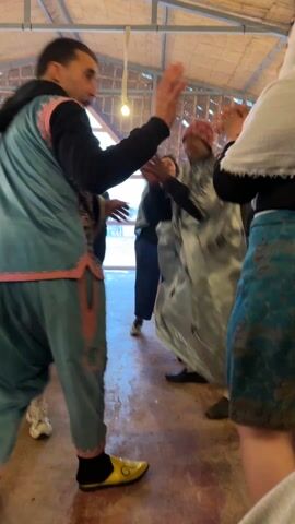 Inside a structure with a thatched roof, a group of people dances to music. A man in a blue and pink outfit, wearing yellow shoes, plays a hand drum. Women in varying attire, including headscarves and colorful skirts, dance around him. Other people watch and clap along. The structure appears to be open-sided, with fencing visible in the background. There's a light bulb hanging from the ceiling.