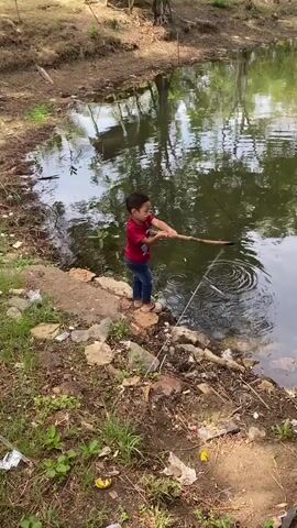 A young child wearing a red shirt and blue jeans stands on the bank of a pond, holding a stick and appearing to fish. The pond's water is green and reflects trees. The bank is covered in dirt, rocks, and some litter. The child's posture suggests they are actively engaged in fishing.