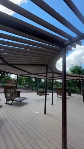 The video pans across a wooden deck area with a slatted canopy overhead. Several wooden swings and benches are positioned on the deck, some appearing empty. In the background, trees, a playground, and a building are visible under a partly cloudy sky. The scene suggests an outdoor recreational space.