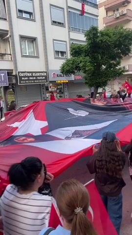 A group of people are marching down a street, holding Turkish flags. They are carrying a large flag featuring a portrait of Mustafa Kemal Atatürk. Some of the marchers are in wheelchairs. Buildings line the street, with businesses such as "ÖZTÜRK ELEKTRİK" and "TERZİ İSMET" visible. A tree is also visible on the side of the street. There is a loud noise in the background.