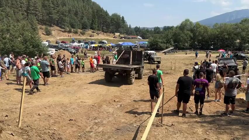 The video shows an outdoor event with many people gathered, watching a truck with a trailer being maneuvered. The ground is dirt and appears to be a designated obstacle course, with some muddy areas and ramps visible. Trees and hills are in the background, indicating a rural or mountainous setting. Several other vehicles, including off-road style trucks and cars, are parked in the background, along with tents and canopies, suggesting a larger gathering or competition. The truck in the foreground is moving, and the trailer it's pulling is tipping at an angle. Spectators are observing the action from behind a barrier. The weather appears to be sunny and warm.