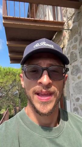 This is a close-up selfie video of a man wearing a dark baseball cap with "St. Barth" embroidered on it and dark-rimmed glasses. He has short brown hair, a beard, and is wearing a green crewneck sweatshirt. The man is speaking and smiling. Behind him, a stone building with a wooden balcony is visible. Further in the background, there are trees and a vineyard with rows of green grapevines stretching down a hillside. The sky is blue with scattered white clouds. The overall scene appears to be outdoors in a rural, possibly Mediterranean setting.