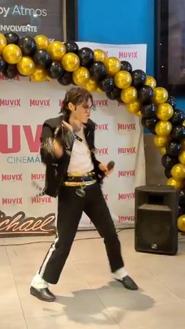 The video shows a person dressed as Michael Jackson dancing in front of a banner that says "MICHAEL JACKSON." The dancer is wearing a black jacket with gold accents, black pants with white stripes, and white socks. The dancer is holding a microphone and performing various dance moves. In the background, there are black and gold balloons arranged in an arch. There is also a speaker on the right side of the frame and a few people sitting and watching the performance. The environment appears to be an indoor public space, possibly a cinema lobby.