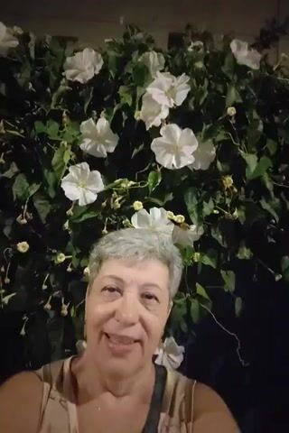 A woman with short grey hair and a light-colored top speaks directly to the camera, smiling, with a lush green plant with white flowers behind her.