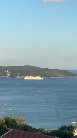 The video shows a large white cruise ship sailing on a blue body of water. In the background, there is a green, hilly coastline with some buildings. The sky is clear and blue. The camera appears to be positioned on a higher vantage point, possibly a hill or balcony, overlooking the scene. The overall impression is one of a serene coastal view with a prominent cruise ship.