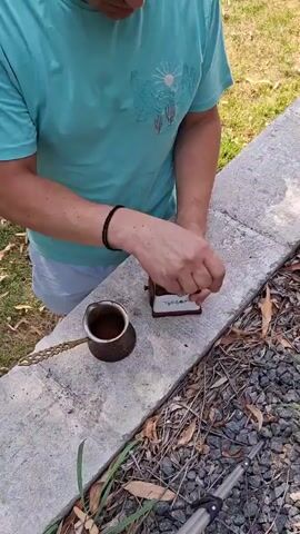 A person wearing a turquoise t-shirt and white shorts is standing outdoors, preparing coffee. They are using a manual coffee grinder, holding it with their left hand and turning the crank with their right. Next to the grinder is a small copper pot with a handle, containing some dark liquid, likely brewed coffee. The person is in the process of transferring ground coffee from the grinder into the pot using a small scoop or spoon. The scene is set on a concrete ledge, with dry leaves and gravel on the ground below. The background shows greenery and a hint of a sunny day.