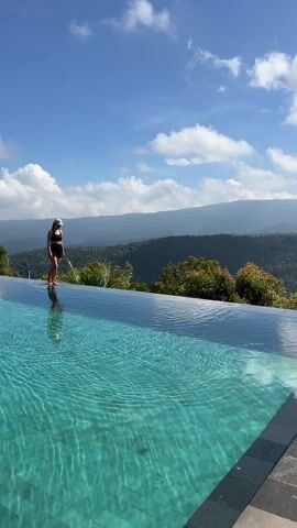 A young woman walks along the edge of an infinity pool against a beautiful mountain backdrop under a bright, blue sky.  She is wearing a black swimsuit and a visor. She stops briefly to kick her leg out, sending water droplets into the air, and then lifts her arms in the air, enjoying the view. The pool is filled with clear, turquoise water that reflects the sky and surrounding mountains. The distant mountains are green, and the sky has scattered white clouds.  The scene is serene and picturesque.