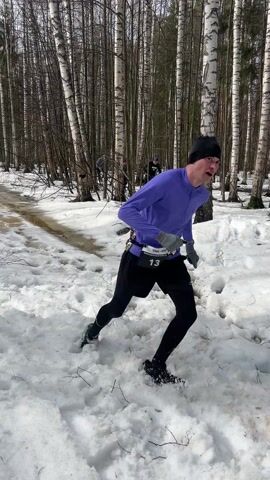 The video shows a snowy birch forest in daylight. Three people are visible, all dressed for cold weather activity. One is running toward the camera in the foreground while the others trail behind. The ground is covered in patchy snow with several deep footprints. There is a shallow pool of standing water to the left of the frame. The runner is wearing a purple long-sleeved shirt, black shorts, and black tights, with athletic accessories at the waist.  The other two runners are further away and are wearing dark clothing and hats. The trees are tall and slender with distinctive white bark.