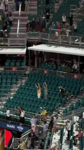 The video shows sections of a stadium with green seating. Some people are standing and waving their arms in the air, while others are walking or sitting. A section of the stadium is labeled "Mari." There are multiple levels of seating. It is daytime.