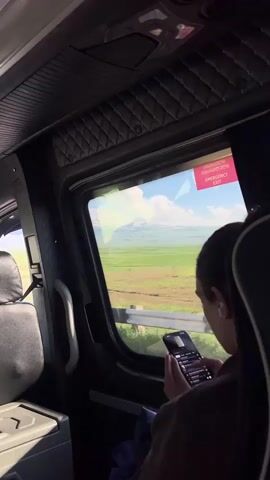 The video shows a person sitting inside a bus, looking at their smartphone. The person is wearing a brown jacket and has earbuds in. Through the window, a vast green field with a distant mountain range under a blue sky with white clouds is visible. A red sign on the window reads "EMERGENCY EXIT" in English and Georgian. The interior of the bus has quilted lining on the ceiling and a grey seat in the foreground. The scene appears to be taking place during the daytime, with bright sunlight illuminating the landscape. The overall mood is peaceful and contemplative, suggesting a journey through a scenic area.