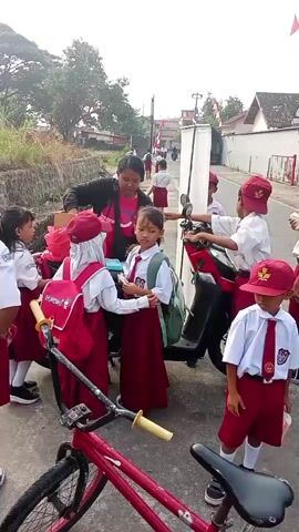 A group of young children wearing school uniforms stand near a motorbike and a bicycle on a paved road. Some of the children have backpacks. One adult, likely a teacher, interacts with the children. A red bicycle is in the foreground. The scene appears to be outdoors, with trees and buildings visible in the background.
