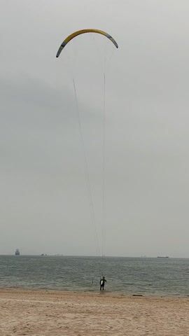 The video shows a person on a beach, standing in shallow water and holding onto the lines of a large kite. The kite is arched in the sky and has a yellow and dark blue or black design. The beach is sandy, and the water appears calm. Several boats are visible in the distance. The sky is overcast.