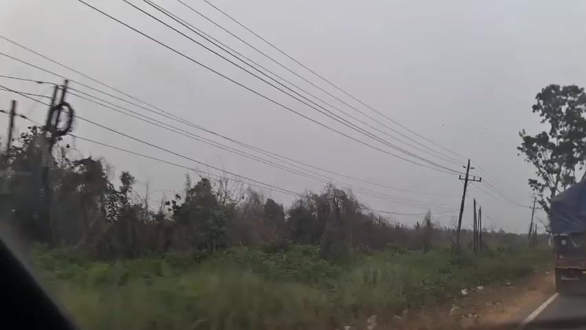 The video shows a view from a moving vehicle, likely a car, on a cloudy day. The camera is focused on the landscape outside the vehicle. The primary elements visible are a series of utility poles with multiple wires running between them, stretching across the frame. To the left of the frame, there is dense vegetation with some trees. In the middle ground, more utility poles and vegetation are visible. To the right of the frame, a large tree is prominent, and further to the right, a dark-colored truck or trailer is partially visible on the side of the road. The road itself is visible in the foreground, appearing to be unpaved or a dirt road. The overall atmosphere is subdued due to the overcast sky. The movement of the vehicle causes a slight blur and a sense of speed.