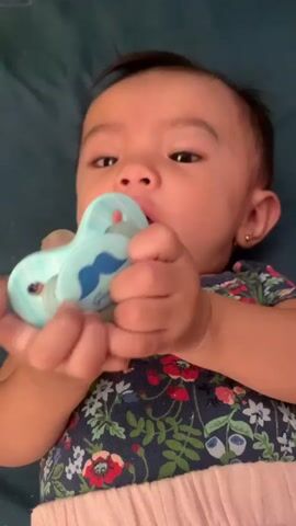 A baby lies on their back, holding a blue pacifier with a mustache design. The baby looks at the camera with its mouth slightly open. The background is a dark blue surface. The baby wears a dark blue shirt with a floral pattern and pink pants.