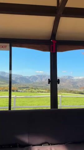 From inside a sheltered structure, the video showcases a picturesque view of a valley, mountains, and trees through a set of windows. The structure has a dark-colored frame and ceiling, and a gray cushioned seating area is visible at the bottom of the frame. A bright red emergency tool hangs on the frame between the windows. Outside, a vibrant green lawn stretches towards the backdrop of rolling mountains beneath a clear, blue sky. An autumn tree with yellow and brown leaves is visible in the right side of the frame. The camera appears to be stationary. Faint conversation can be heard in the background.