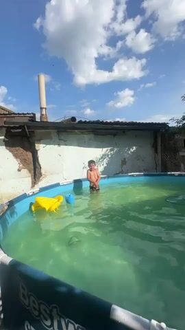 A young boy wearing dark swim trunks stands in a medium-sized, above-ground swimming pool. The pool water is a murky green color. A yellow inflatable duck toy floats on the left side of the pool. Behind the pool is a dilapidated building with a rough, light-colored wall and a corrugated metal roof. A tall, cylindrical chimney or pipe is visible extending from the roof. The sky above is bright blue with scattered white clouds. The boy is partially submerged in the water, with his upper body and head visible. He appears to be looking around the pool.