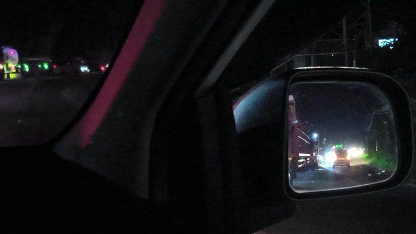 The video shows the interior of a vehicle at night or in a very dark environment, possibly a tunnel. The primary focus is on the side mirror, which reflects a scene behind and to the side of the vehicle. In the reflection, multiple vehicles, likely large trucks or buses, are visible, illuminated by their own headlights and numerous flashing lights (red, blue, and green). These lights suggest an emergency situation or a special convoy. The vehicles appear to be within a confined space, possibly a tunnel, indicated by the continuous structure above and to the sides of the reflected roadway.

Through the passenger-side window, distant flashing lights, predominantly red and green, are also visible against the dark backdrop, reinforcing the presence of other vehicles. The interior of the recording vehicle is dark, with only faint reflections and contours of the door frame and pillar being discernible. There are no visible people or text. The location context is Keudetanjong, Indonesia.