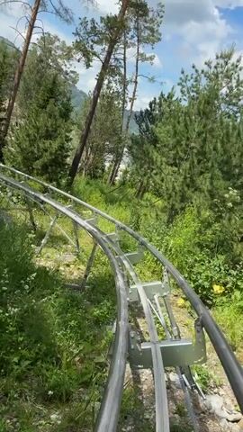 The video shows a first-person perspective of someone riding a roller coaster through a forest. The roller coaster tracks are made of metal and are supported by metal beams. The surrounding environment is lush and green, with tall trees and vegetation. The sky is blue with some white clouds. The person's feet are visible at the bottom of the frame, wearing sandals. The roller coaster appears to be moving at a moderate speed, going around curves and down gentle slopes. In the distance, there are glimpses of mountains and water. There are other people visible on the roller coaster in the distance.