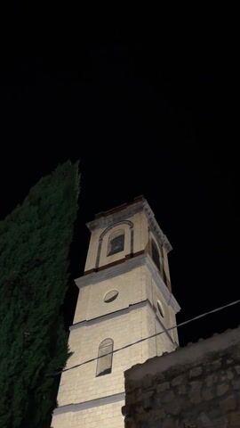 The video shows a low-angle view of a church bell tower at night. The tower is made of light-colored stone and has several arched windows. A tall, dark green tree stands to the left of the tower. The sky is black, and the overall lighting is dim. The sound of church bells can be heard. A stone wall is visible in the lower right corner of the frame, partially obscured by shadows. A thin wire or cable stretches across the lower portion of the frame.