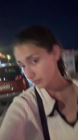 A woman with dark hair pulled back is filmed at night, looking at the camera and slightly away from it. She is wearing a white collared shirt with black trim. The background is dark and out of focus, with some lights and a hint of a car visible. The video appears to be a selfie taken outdoors at night.