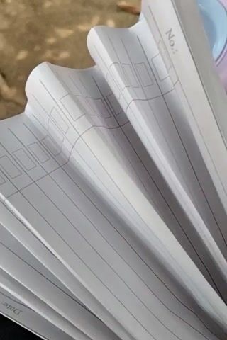 The video shows a close-up, Dutch angle shot of a stack of papers. The papers are lined and have boxes for entries, suggesting they are from a ledger or a form. In the upper left corner, text that appears to say "No:" is visible, along with small empty boxes below it. The pages are fanned out and appear to be slightly curled or wavy. The background consists of a blurred, earthy texture, possibly ground or dirt, and a portion of a blue and pink object that could be a toy or a container. The overall lighting suggests an outdoor or well-lit indoor environment.