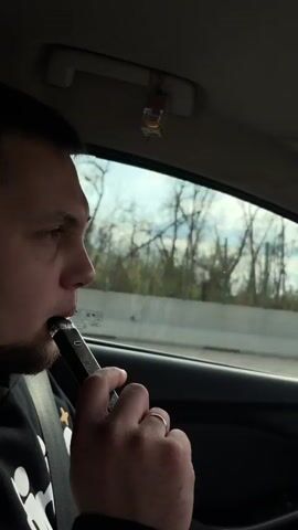 A man is seen from the side in the driver's seat of a car. He is holding a vape pen to his lips, inhaling and exhaling a large cloud of vapor. He has short dark hair and a trimmed beard. He is wearing a black hoodie. He is wearing a seatbelt. A wedding band is visible on his left ring finger. A small, possibly decorative object hangs from the car's ceiling.  The window behind him shows trees and a concrete barrier, suggesting he is on a highway. The car's interior is dark.