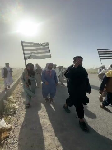 The video captures a group of men walking outdoors in what appears to be a march or procession in Pishin, Pakistan. The scene is brightly lit by the sun, indicating daytime, and the environment is dusty and arid.

In the opening shot, a young person, appearing male, leads the group while carrying a large flag with horizontal black and white stripes on a wooden pole. Other men, dressed in traditional South Asian attire such as shalwar kameez, tunics, vests, and various head coverings or scarves, follow behind.

Subsequent close-up shots feature different individuals walking towards the camera:
- A young man in a white tunic and trousers, a brown vest, and a white scarf, walks briskly with a determined expression.
- A group of men, some smiling, walk together. One man wears a light blue tunic, blue trousers, and a white shawl, while another wears a light brown tunic and a maroon scarf.
- A man in a light blue tunic and a black baseball cap with "NYC" written on it also walks towards the camera, smiling.

The group moves along a dirt path or road adjacent to an asphalt road. The bright sun creates strong shadows and lens flare throughout the video. The overall mood appears calm and collective, with individuals showing a mix of determined expressions and smiles.