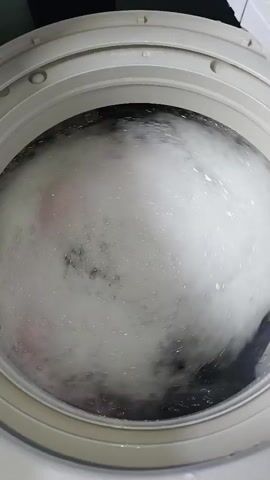 The video shows a close-up, top-down view of the inside of a washing machine during a wash cycle. The drum is filled with soapy water and a load of laundry. The clothes, which appear to be a mix of dark and light colors, are being agitated and spun by the machine, creating a swirling vortex of suds and water. The focus is on the movement of the clothes and the foam, which obscures most of the details of the laundry itself. The washing machine appears to be a top-loading model, indicated by the open lid at the top of the frame.
