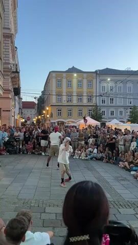 Two men are street performing in a cobblestone plaza in front of a large crowd. The man on the left is wearing a white t-shirt and khaki shorts and is dancing. The man on the right is wearing a white t-shirt and khaki shorts and is also dancing. They are surrounded by a crowd of people who are sitting and standing to watch them perform. Behind the crowd are buildings with yellow and grey facades and a clear blue sky above.