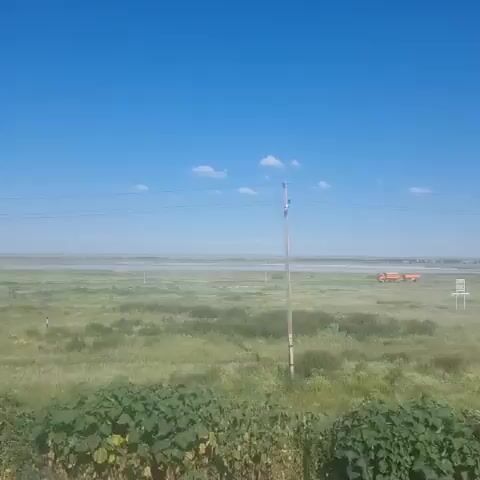 The video shows a landscape with a clear blue sky dotted with a few white clouds. A vast, green, grassy field stretches out towards a distant horizon, where a faint body of water or a haze can be seen. In the middle ground, there is a red structure that appears to be a small building or vehicle, possibly part of an airport or industrial facility, along with some white posts. In the foreground, there is a dirt path or road. A single utility pole with wires is visible on the left side of the frame. The overall scene suggests an open, possibly rural or undeveloped area under a bright, sunny sky.