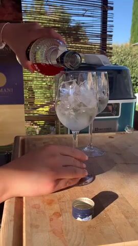 A person's hand is seen pouring a dark red liquid from a clear glass bottle with a dark cap into a wine glass filled with ice. Another wine glass is visible behind it. In the background, there is a blue ice maker and some foliage. A wooden cutting board is in the foreground with a bottle cap on it.