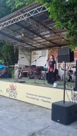 A medium full shot from a slightly low angle captures a woman singing on a stage outdoors. The stage has a corrugated metal roof and exposed metal framing. The woman is in the center, wearing a black top and a long black skirt with red embroidery or patterns, holding a microphone. To her left is an old wooden cart, and to her right are drum sets and various audio equipment. A large black speaker is visible in the foreground on the right. A yellow banner with text is stretched across the front of the stage. Trees are visible in the background and to the left and right of the stage. The ground is paved.