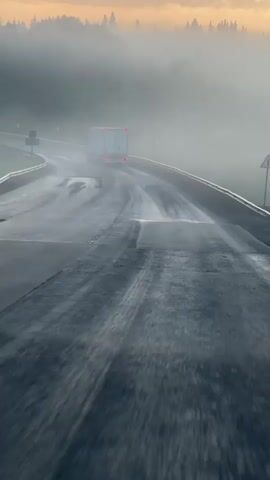 A semi-truck drives on a wet road through a foggy forest. The sun is setting, casting an orange glow on the clouds. The road is curved and has guardrails on the sides. The truck is white and has its brake lights on. The fog is thick, obscuring the trees in the background. The video is filmed from a moving vehicle, indicated by the blurred foreground.