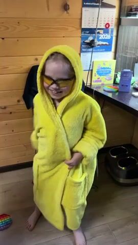 A young child in a yellow hooded bathrobe and yellow-tinted sunglasses is dancing in a room with wooden paneling. The child is barefoot and has a wide smile. A calendar, a black chair, and a foot massager are visible in the background. A colorful fidget toy is on the floor.