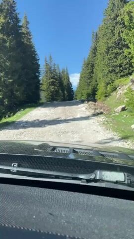 The video shows a view from inside a vehicle driving on a gravel road that ascends through a forest. The road is covered in loose stones and dirt, and is flanked by tall evergreen trees on both sides. The sky is clear and blue. The camera is positioned to show the dashboard of the vehicle and the road ahead. The video is approximately 4 seconds long.