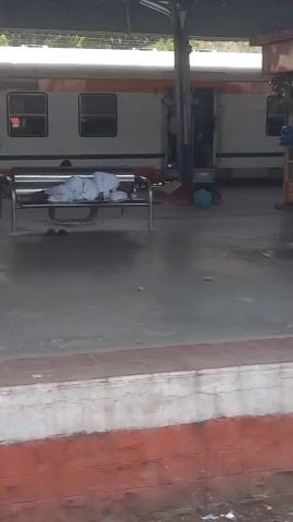 The camera moves from left to right along a train platform. A tea stall with beverages is seen, along with several passengers waiting on the platform, and a train alongside the platform. The platform itself has a red and white edge and concrete floor. There are metal pillars supporting the platform roof.