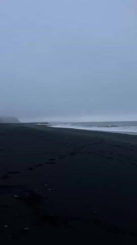 The video shows a black sand beach on a cloudy day.  Waves are breaking on the shore.  There are footprints in the sand.  The sky is overcast and grey. The water is also grey and choppy. The coastline is visible in the distance, obscured by mist or fog.