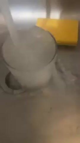 The video shows a glass mug being filled with water in a stainless steel sink. The water overflows as it fills, and there is a yellow sponge next to the mug. The sink drain is visible.