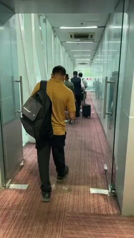 A man wearing a yellow shirt and black pants walks down a jet bridge carrying a black backpack. He is followed by other people with luggage. The jet bridge is enclosed by glass on the right side and white walls on the left side. The floor is covered with a red carpet. Fluorescent lights are visible on the ceiling.