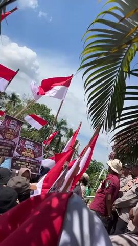 The video shows a gathering of people outdoors under a bright, cloudy sky. Many individuals are holding up Indonesian flags, which are red and white. Some people are wearing uniforms that appear to be those of law enforcement or security personnel. In the background, there are banners with text and images, suggesting a planned event or demonstration. A group of people are standing on top of a platform or vehicle, with one person speaking into a microphone, possibly addressing the crowd. Large palm fronds are visible in the foreground, partially obscuring the view. The overall atmosphere suggests a public assembly or rally.