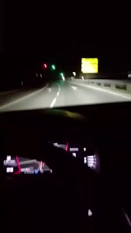 The video depicts a nighttime scene from inside a car driving on a road. The road has white lane markings and is illuminated by the car's headlights. In the distance, traffic lights are visible, some displaying red and others green. To the right of the road, there's a sign, though it appears blurry and illegible. The car's dashboard is visible in the foreground, displaying digital information. The lighting is predominantly dark, with the exception of the bright headlights and the illuminated dashboard and traffic signals.