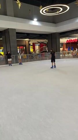 This is a video taken from an ice skating rink, likely indoors due to the ceiling structure and lighting. Several people are visible on the ice, wearing skates. One person, wearing a black outfit, is skating in the foreground. In the background, another person, in a light blue top and white shorts, is also skating. Further left, another person in a black and white outfit is standing near the edge of the rink. The ice surface appears smooth, and there is a barrier around the rink. Beyond the barrier, stores or food establishments can be seen, suggesting the rink is located in a mall or entertainment complex. Decorative star-shaped ornaments and circular light fixtures hang from the ceiling. The lighting is bright, illuminating the entire scene.