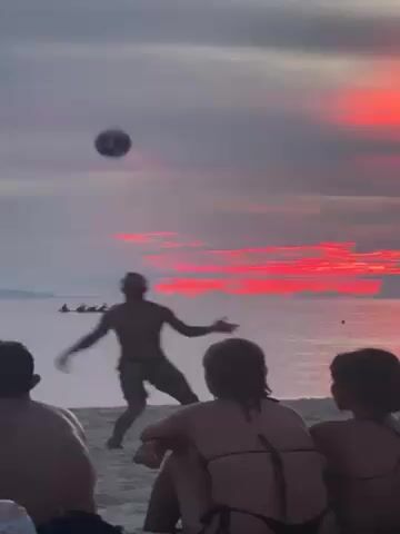Several people are relaxing on a beach, silhouetted against a red and grey sunset sky. Two young women are sitting with their backs to the camera, wearing bikini tops.  A bare-chested man with dreadlocks is playing with a ball on the sand. Another bare-chested man runs in the background. In the distance, a boat with several people in it can be seen on the water, silhouetted against the bright sky.