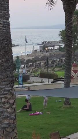 A vertical video shows a seaside resort area. In the foreground, two palm trees frame the view. On a grassy area, a woman is sitting on a pink yoga mat, facing away from the camera. A child in a white shirt is standing and facing the woman. To the right of the woman and child, a younger child in a pink top is running. In the background, there is a beach with lounge chairs and umbrellas, a Greek flag flying, a statue of a person, a building that appears to be a restaurant or bar, and a small red and gold building resembling a chapel. The sky is overcast.