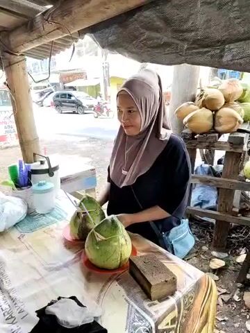 A woman wearing a hijab and a black shirt stands behind a table, in what appears to be a small outdoor market stall. On the table are two green coconuts, each with a straw inserted, sitting on red plates. There is also a dark brown, rectangular object, possibly a block of something, near the coconuts. Behind the table is a small wooden shelf stacked with more coconuts. The stall is covered by a simple roof structure supported by wooden posts. In the background, there is a road with cars and motorcycles passing by, and some buildings are visible. The woman points to the side of the table.