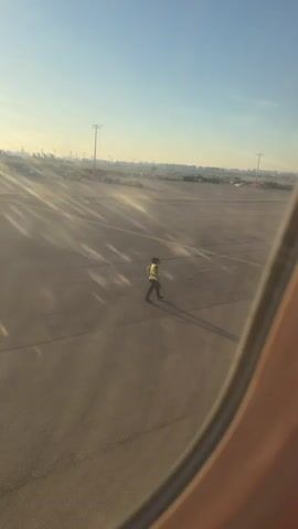 The video captures a person on an airport tarmac, seen through an airplane window. The sun is low in the sky, casting long shadows. The person is wearing a reflective vest and appears to be walking. The tarmac is paved and marked with lines. In the distance, there are utility poles and what appear to be parked vehicles or aircraft. The sky is clear and blue.