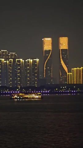 This media file depicts a nighttime scene of a cityscape, likely in Shangcheng District, China. The dominant subjects are modern skyscrapers illuminated with golden lights, creating a striking visual against the dark sky. Two particularly distinctive twin towers in the center feature a twisting, ribbon-like lighting design that accentuates their architectural form. To the left and right of these towers are rows of other tall buildings, also lit up, contributing to a vibrant, urban panorama.

In the foreground, a ferryboat or tour boat is visible traversing a body of water. The boat is also illuminated, with lights visible through its windows and along its deck, suggesting it is carrying passengers. The water reflects some of the city lights, adding depth to the scene. The overall atmosphere is one of nighttime urban activity and spectacle. The weather appears clear, allowing for a sharp view of the illuminated cityscape. The time of day is definitively night, indicated by the darkness of the sky and the prominence of artificial lighting. There is no visible text in the media file.